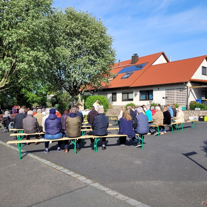 Mehrere Menschen sitzen auf Bierbänken unter einem Baum und feiern Gottesdienst.