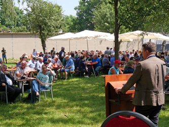 Man sieht den Redner Kirchner von hinten und schaut auf die Zuhörer. Sie sitzen im Biergarten.