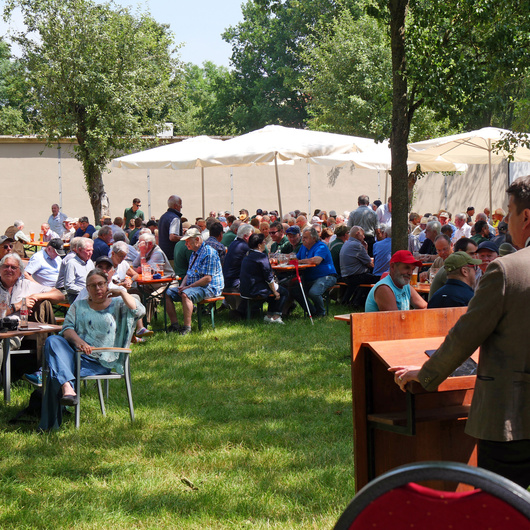 Man sieht den Redner Kirchner von hinten und schaut auf die Zuhörer. Sie sitzen im Biergarten.