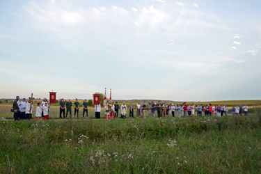 Das ist ein Blick in die Landschaft mit einer Blumenwiese im Vordergrund. Hinten stehen in einer Reihe Menschen und beten oder singen. Sie sind recht klein zu sehen.