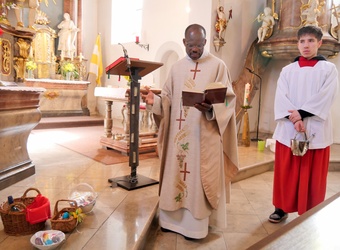 Das Bild zeigt eine kirchliche Zeremonie. Ein Priester in weißer Robe mit goldenen Verzierungen und einem Kreuz steht an einem Lesepult und liest aus einem Buch. Neben ihm steht ein junger Mann in weißer Albe mit rotem Gürtel und hält einen Weihrauchkessel. Im Vordergrund befinden sich bunte Ostereier in Körbchen.