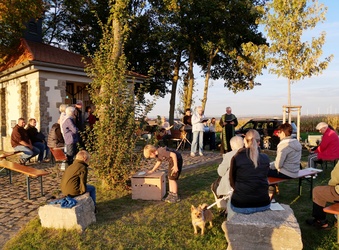 Das Bild zeigt eine Kapelle im Freien im Licht dre Abendsonne. Sie ist links im Bild. Rechts sitzen Menschen auf Steinen und Bänken. Ein kleiner Hund ist in der Mitte.