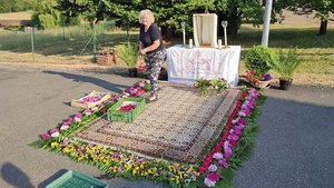 Eine Frau geht auf einem Teppich, der von Blumen umrahmt ist. Im Hintergrund ist ein Altar aufgebaut.