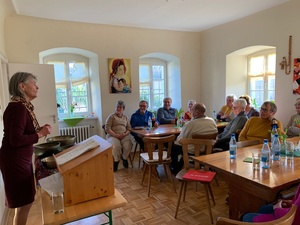 Links im Bild steht eine Frau an einem Tisch, auf dem mehrere Klangschalen stehen, und erzählt etwas. Rechts im Bild sieht man mehrere Leute, die an Tischen sitzen und aufmerksam zuhören.