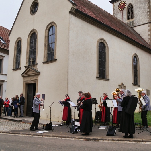 Eine Musikkapelle in ihrer Tracht spielt. Im Hintergrund ist die Hausener Kirche zu sehen.