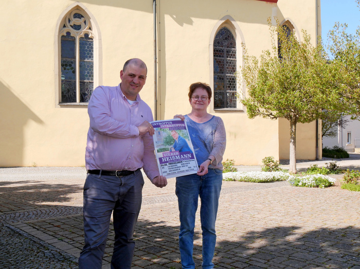 Das Bild zeigt zwei Personen, die vor einer Kirche mit großen Bogenfenstern stehen. Der Mann links trägt ein lila Hemd und dunkle Hosen, die Frau rechts trägt ein graues Oberteil und Jeans. Sie halten gemeinsam ein Plakat, auf dem ein Konzert mit Volker Heißmann beworben wird.