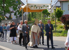 Auf dem Bild sieht man eine Fronleichnamsprozession. Geistliche und Gemeindemitglieder tragen ein Baldachin, begleitet von Zuschauern in einer blumengeschmückten Straße.