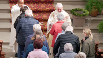 Zwei Priester segnen ältere Menschen. Die Priester sind von vorn zu sehen, die älteren Menschen von hinten.