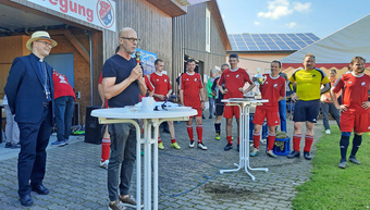 An einem weißen Stehtisch steht der Moderator mit einem Mikrofon in der Hand. Links hinter ihm steht Bischof Franz Jung.