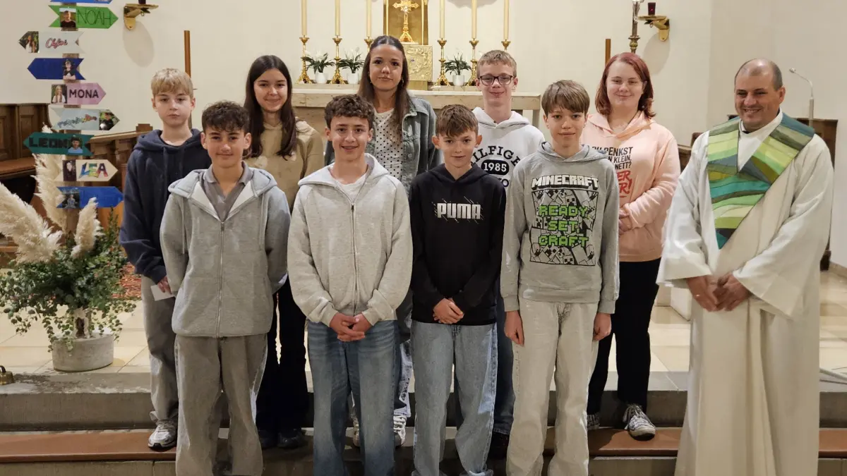 Das ist ein schönes Gruppenbild mit zehn Kindern. Es ist in einer Kirche aufgenommen worden. Rechts auf dem Foto ist der Pastoralreferent.