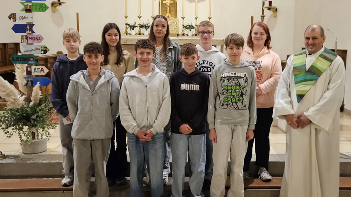 Das ist ein schönes Gruppenbild mit zehn Kindern. Es ist in einer Kirche aufgenommen worden. Rechts auf dem Foto ist der Pastoralreferent.