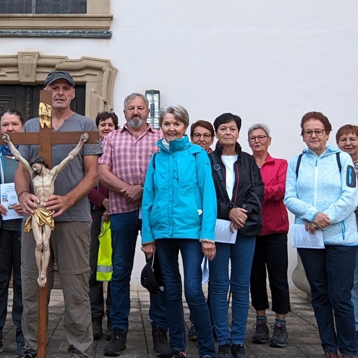 Eine Gruppe von 13 Menschen steht vor einer Kirche. Ein Mann hält vor sich ein großes Kreuz.