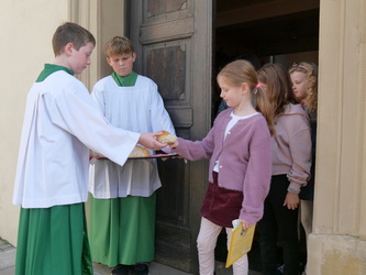 Auf dem Bild sind fünf Kinder zu sehen. Die zwei Ministranten stehen mit einem Tablet mit Brot vor einer Kirchentüre. Die Mädchen rechts bekommen Brotstücke in ihre Hände.