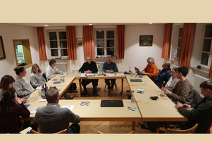 Das Bild zeigt eine Gruppe von Menschen, die an einem rechteckigen Tisch in einem hellen Raum mit Fenstern und Vorhängen sitzen. Sie nehmen an einer Besprechung teil, mit Papieren und Getränken auf dem Tisch. Die Atmosphäre wirkt formell und konzentriert.