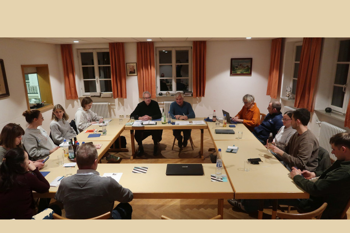 Das Bild zeigt eine Gruppe von Menschen, die an einem rechteckigen Tisch in einem hellen Raum mit Fenstern und Vorhängen sitzen. Sie nehmen an einer Besprechung teil, mit Papieren und Getränken auf dem Tisch. Die Atmosphäre wirkt formell und konzentriert.