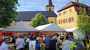 Im Vordergund sind Musikanten. Hinter ihnen sind weiße Zeltdächer, unter denen sitzende Menschen zu sehen sind. Im Hintergrund ist eine Kirche.