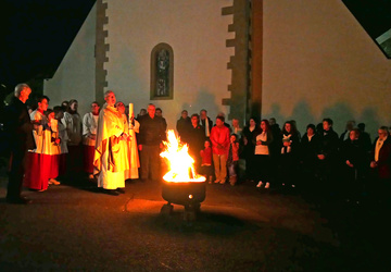 Ein Bild mit mehreren Menschen im Dunkeln vor der Kirche. In der Mitte brennt das Osterfeuer. 