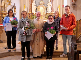 Das Bild zeigt sechs Personen in einer Kirche. Im Mittelpunkt steht ein Priester in liturgischer Kleidung, der einen Blumentopf hält. Die anderen Personen halten Blumen und Zertifikate, während sie neben dem Priester stehen und lächeln. Im Hintergrund sind Statuen und eine goldene Altardekoration sichtbar.