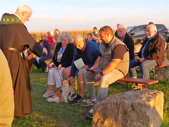 Das ist ein stimmungsvolles Bild in rötlicher Abendsonne. Links steht ein Pfarrer und segnet. Rechts sind eine Gruppe Menschen und ein paar Hunde.