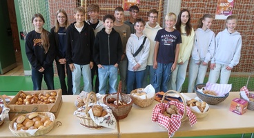 Das Bild zeigt mehrere Kinder, die hinter einem Tisch stehen. Sie schauen in die Kamera. Auf dem Tischliegen Brote und Brötchen in Weidekörbchen.