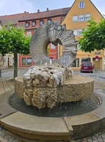 Die Aufnahme zeigt einen steinernen Brunnen mit einer Skulptur, die eine Gruppe von Menschen darstellt. Er steht auf einem gepflasterten Platz in einem dörflichen Umfeld mit bunten Häusern.