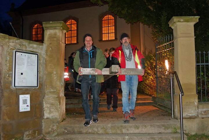 Es ist dunkel. Zwei Männer tragen die Querbalken eines Holzkreuzes. Im Hintergrund sieht man die Krirche St. Vitus mit ihren erleuchteten Fenstern.