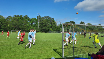 Blick von außen auf das Fußballfeld mit dem Tor auf der rechten Seite. Der Fußball ist hoch in der Luft vor einem blauen Himmel.