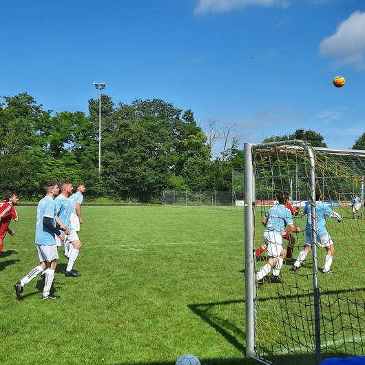 Blick von außen auf das Fußballfeld mit dem Tor auf der rechten Seite. Der Fußball ist hoch in der Luft vor einem blauen Himmel.