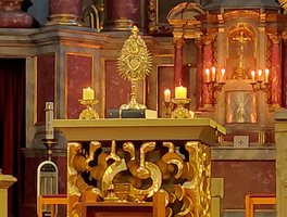 Das Bild zeigt einen vergoldeten Altarbereich in einer Kirche. Im Zentrum steht ein Monstranz mit einem Herzmotiv, flankiert von zwei brennenden Kerzen. Hinter dem Altar sind weitere Kerzen und ein Kreuz zu sehen.