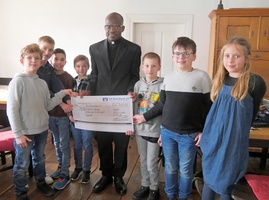 Das Bild zeigt eine Gruppe von sieben Kindern, die zusammen mit einem Priester in einem Raum stehen. Sie halten einen großen Scheck über 300 Euro. Der Raum ist im historischen Rathaus in Rieden.