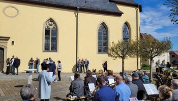 Blick von hinten auf die Blaskapelle, im Hintergrund stehen mehrere Menschen vor der Wand der Kirche und unterhalten sich.