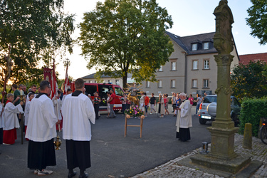 Auf dem Platz vor der Kirche stehen in einem Rund mehrere Menschen. Rechts steht der Pfarrer und liest gerade etwas aus einem Buch vor.