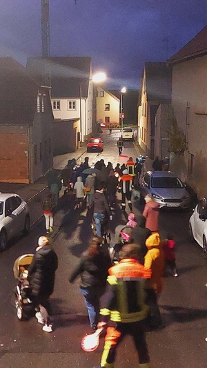 Eine Gruppe Kinder und Erwachsener gehen begleitet von Feuerwehrmännern mit Laternen durch eine Dorfstraße.