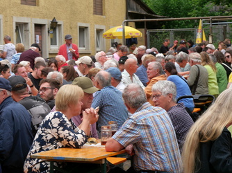 Auf dem Bild sieht man viele Menschen, die draußen an Tischen sitzen, essen, trinken und sich in geselliger Runde unterhalten. Es herrscht eine fröhliche Atmosphäre.