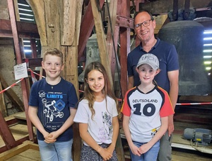 Vor der Glocke im Glockenturm stehen der Kirchenpfleger und die drei Kinder. Sie schauen glücklich in die Kamera.