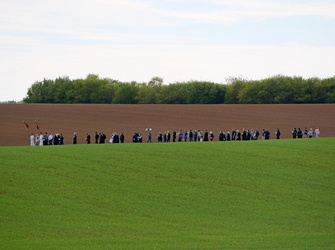 Das Bild zeigt eine Gruppe von Menschen, die sich am Rand eines Feldes versammelt haben. Vor der Wandergruppe ist ein Feld mit grünem Gras, hinter ihnen befindet sich ein frisch gepflügtes Feld und ein Waldstreifen. Einige Personen halten Fahnen oder Schilder. Der Himmel ist klar mit einigen Wolken.