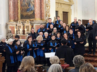 Das Bild zeigt einen Chor, der in einer Kirche auftritt. Die Chormitglieder tragen schwarze Kleidung mit blauen Schals. Sie stehen in mehreren Reihen und halten Notenblätter in den Händen. Im Vordergrund sind Zuschauer zu sehen, die der Darbietung folgen.