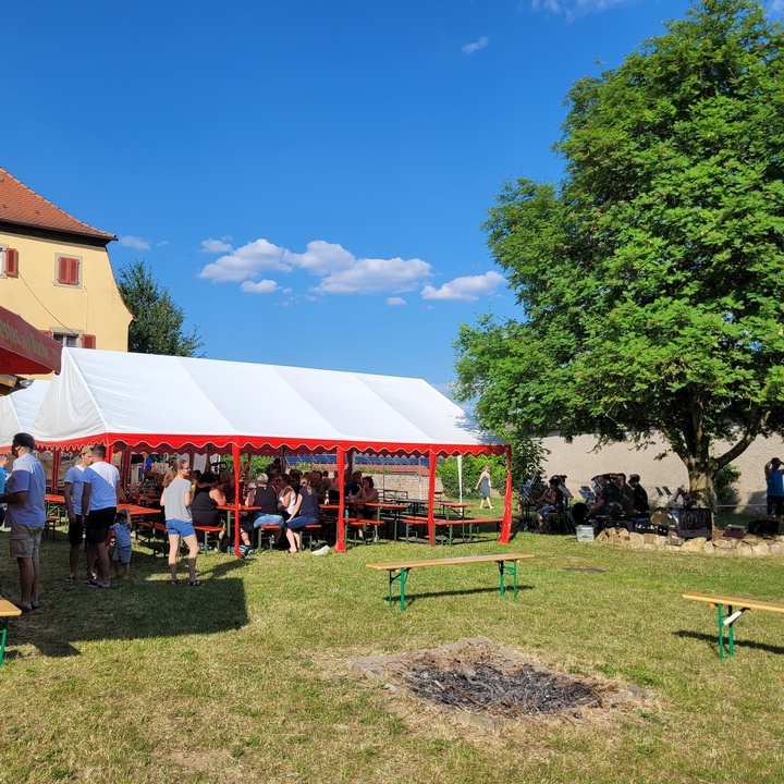 Außenaufnahme des Festgeländes. Im Vordergrund eine Lagerfeuerstelle, im Hintergrund das Festzelt und ein Baum.