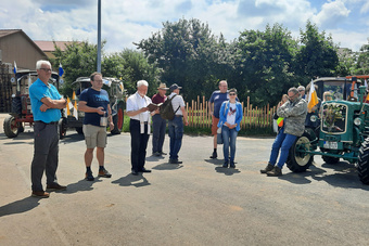 Eine kleine Menschengruppe steht auf dem Parkplatz mit den Traktoren. Pater Matthäus liest aus einem Gebetsbuch vor.