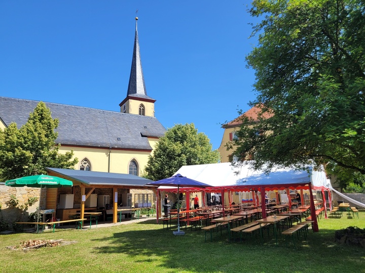 Blick auf einen Pfarrgarten, im Vordergrund sieht man Zelte, unter denen Biertische und -bänke aufgebaut sind, im Hintergrund sieht man die Prosselsheimer Kirche