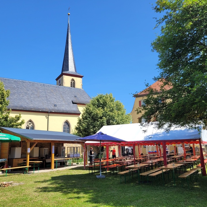 Blick auf einen Pfarrgarten, im Vordergrund sieht man Zelte, unter denen Biertische und -bänke aufgebaut sind, im Hintergrund sieht man die Prosselsheimer Kirche