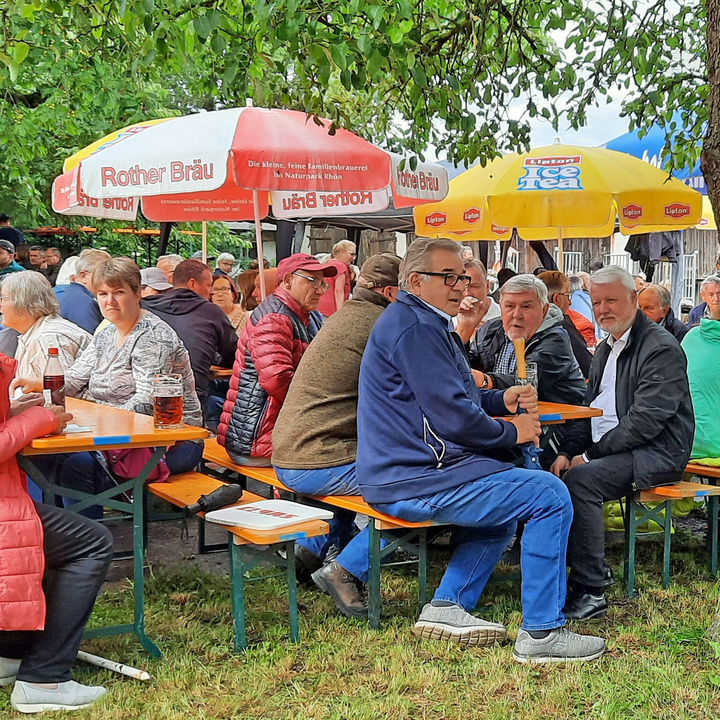 Mehrere Menschen sitzen auf Bierbänken unter grünen Bäumen. Unter ihnen ist Pater Matthäus. Er schaut herüber zur Kamera.