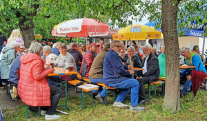 Mehrere Menschen sitzen auf Bierbänken unter grünen Bäumen. Unter ihnen ist Pater Matthäus. Er schaut herüber zur Kamera.