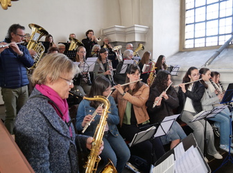 Dieses Bild zeigt ein Orchester, das in einem hellen, hohen Raum spielt. Es sind verschiedene Musiker mit Blasinstrumenten wie Trompeten, Flöten und Saxophonen zu sehen. Die Musiker sitzen auf Stühlen und spielen ihre Instrumente, während sie auf Notenblätter blicken.