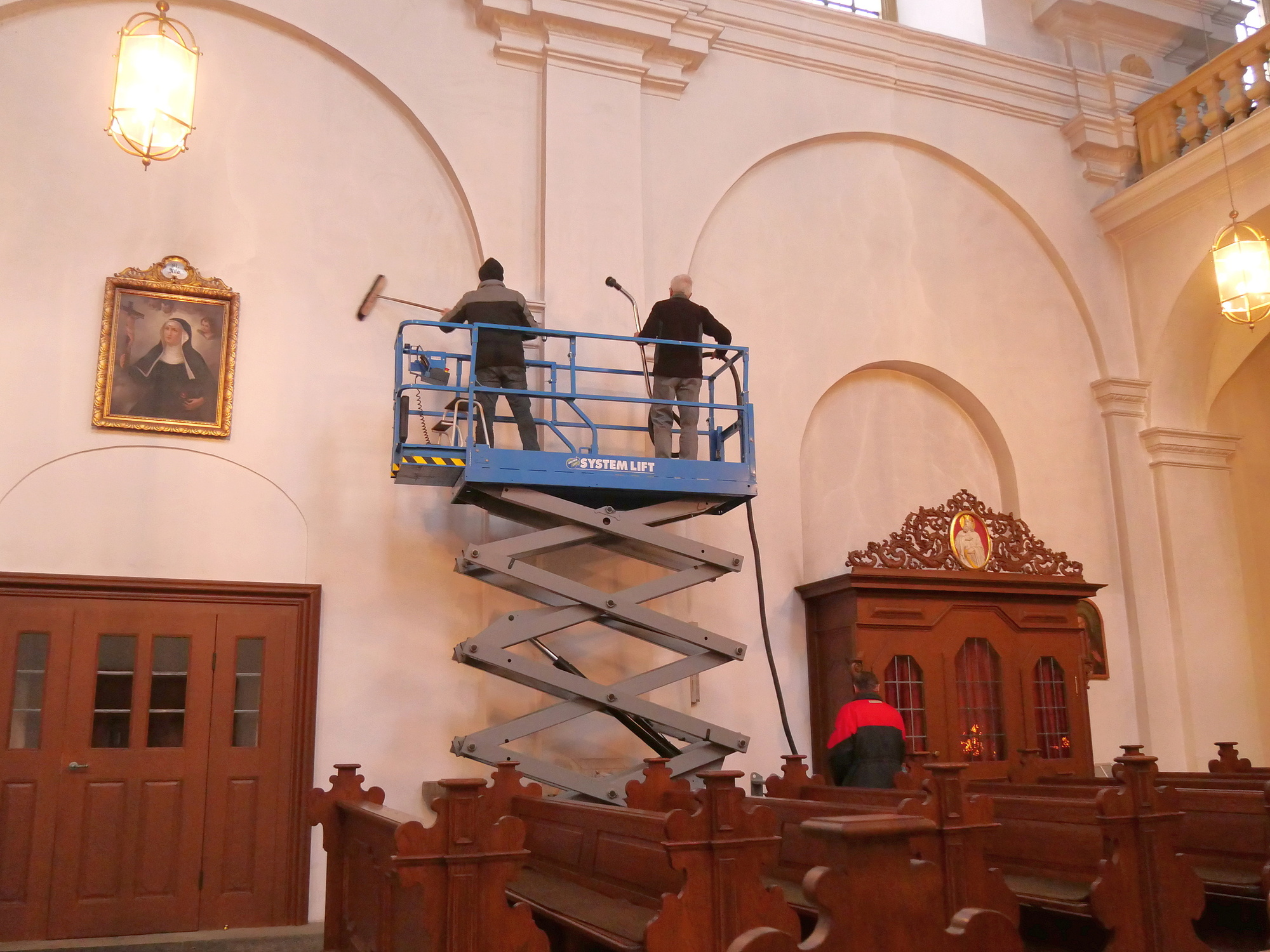 Zwei Männer reinigen mit dem Staubsauger und Besen eine Kirchenwand.
