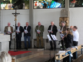 Auf dem Bild stehen mehrere Männer in einer Kirche, einige halten Blätter und singen. Ein kleines Musikensemble spielt im Vordergrund. Ein Kreuz hängt an der Wand.
