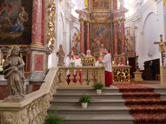 Das Bild zeigt eine festliche Kirche mit einer prunkvollen Barockausstattung. Am Altar stehen ein Priester und mehrere Kinder in liturgischen Gewändern. Statuen und Gemälde schmücken den Hintergrund.