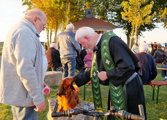 Auf diesem Bild sieht man im Hintergrund eine kleine Kapelle im Abendlicht. Vorne streichelt ein Pater einen kleinen Hund, der auf einem Stein sitzt. Rechts im Bild ist offenbar der Hundebesitzer. Er schaut freundlich.