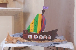 Ein gebasteltes Schiff mit bunten Segeln aus Papier. Auf dem Schiff sind Fotos von Kindergesichtern aufgeklebt.