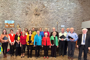 Das Bild ist ein Gruppenbild mit den Chormitgliedern und dem Chorleiter. Es ist in einer Kirche fotografiert worden. Alle freuen sich sichtlich.
